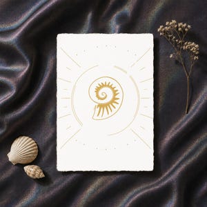 Peut inclure: Une carte rectangulaire blanche aux bords effilochés présentant une illustration au trait doré d'un coquillage en spirale centré dans un cadre circulaire avec des rayons de soleil, accompagnée de deux petits coquillages et d'une fleur séchée.