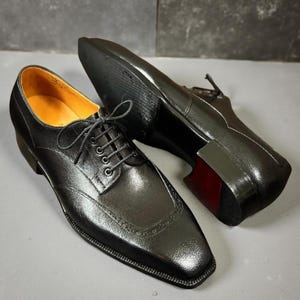Peut inclure: Une paire de chaussures de ville en cuir noir avec un bout carré et une fermeture à lacets. Les chaussures ont une finition texturée et un accent de semelle rouge. L'intérieur de la chaussure est de couleur beige clair.