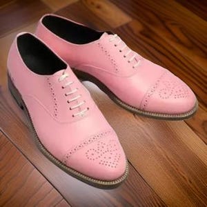 May include: A pair of pink leather Oxford shoes with white laces. The shoes feature a classic design with decorative perforations on the toe and sides. The shoes are set on a wooden surface.