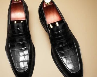 Peut inclure: Paire de mocassins en cuir noir avec une texture peau de crocodile. Les chaussures ont un design classique avec un bout arrondi et un petit talon. L'intérieur est doublé d'un matériau marron clair, et les embauchoirs sont visibles.
