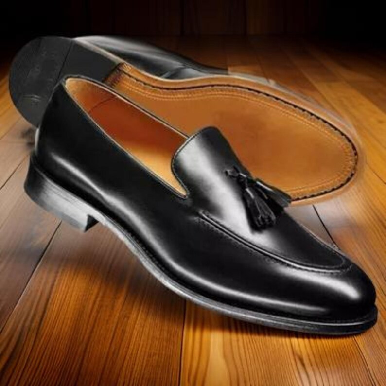 May include: A pair of black leather loafers with a tassel detail. The shoes have a classic design with a rounded toe and a low heel. The sole of one shoe is visible in the background, showing a light brown color.
