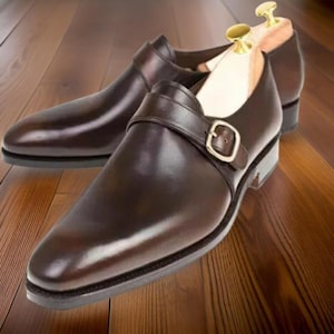 Könnte beinhalten: Ein Paar braune Monk-Schuhe aus Leder mit einer einzigen Schnalle an jedem Schuh. Die Schuhe befinden sich auf einer Holzoberfläche.