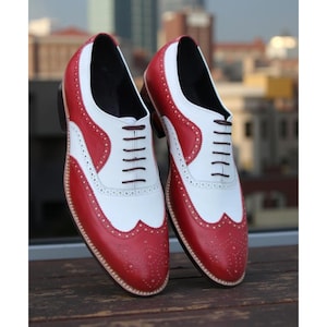 May include: A pair of red and white leather Oxford shoes with a classic wingtip design. The shoes feature a perforated pattern along the edges and a brown sole. The laces are dark brown, and the shoes are displayed on a wooden surface.