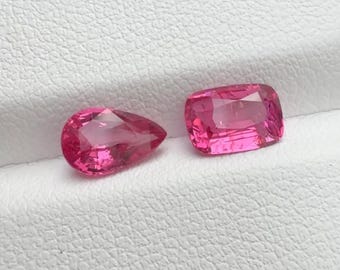 May include: Two vibrant pink gemstones are displayed side by side. One gemstone is cut into a pear shape, while the other is cut into a rectangular cushion shape. Both stones feature faceted surfaces that reflect light, showcasing a deep, saturated pink hue.