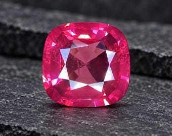 May include: A cushion-cut gemstone with a vibrant, saturated pinkish-red hue. The stone features a faceted surface that creates a star-like pattern of light reflections across its top, showcasing high clarity and a brilliant, polished finish.