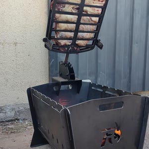Puede incluir: Una parrilla de carb&oacute;n de metal negro con una rejilla vertical ajustable que sostiene un trozo grande de carne cruda. La base de la parrilla tiene un dise&ntilde;o recortado de una cabeza de toro con llamas visibles en el interior.