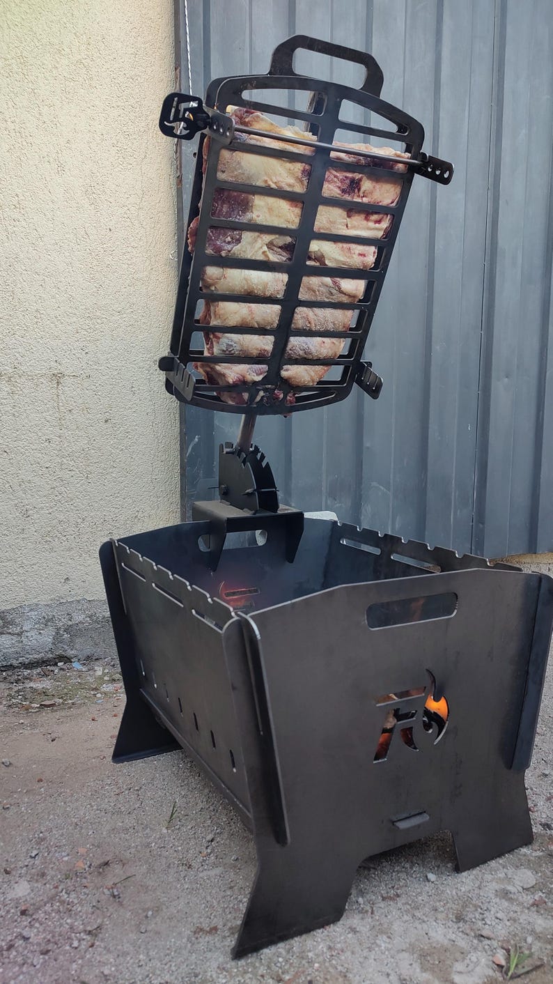 Puede incluir: Una parrilla de carb&oacute;n de metal negro con una rejilla vertical ajustable que sostiene un trozo grande de carne cruda. La base de la parrilla tiene un dise&ntilde;o recortado de una cabeza de toro con llamas visibles en el interior.