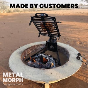 Puede incluir: Una parrilla de metal negro ajustable montada sobre una base circular de color claro para fogata con textura de placa de diamante. La parrilla sostiene alimentos sobre un fuego de le&ntilde;a. Texto superior MADE BY CUSTOMERS e inferior METAL MORPH Design in metal.