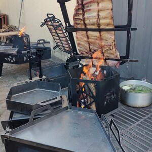 Puede incluir: Un sistema de parrilla exterior de metal negro con un soporte vertical que sostiene un costillar sobre fuego abierto, una plancha triangular plana, una caja de fuego hexagonal, una rejilla de cocci&oacute;n y una olla plateada con hierbas.