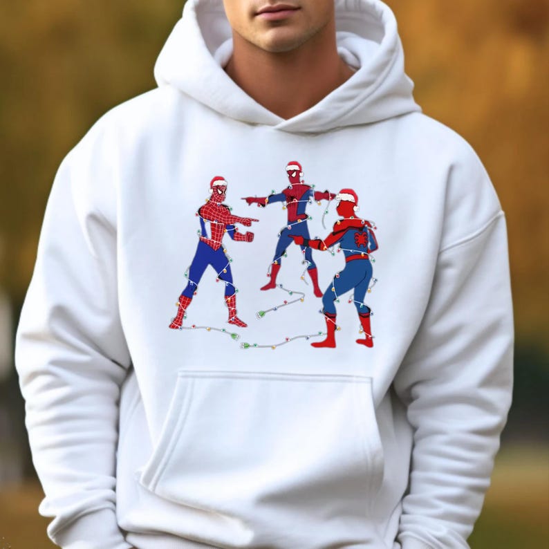 May include: White hooded sweatshirt featuring a graphic of three Spider-Man figures in Christmas hats, entangled in string lights. The figures are in red and blue costumes, with one pointing. A festive holiday design.