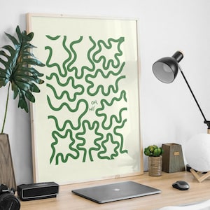 May include: A framed print with a green abstract design on a cream background. The design features wavy lines and the text "OH, HI!" in the center. The print is hanging on a white wall above a desk with a laptop, a lamp, and a plant.