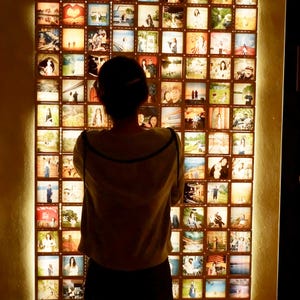 May include: A large, rectangular wall display composed of numerous illuminated square panels, each featuring a unique photograph. A person stands with their back to the camera, facing the grid of backlit images that create a warm, glowing effect.