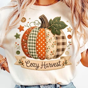 May include: A cream-colored t-shirt featuring a large graphic of a patchwork pumpkin with various patterns including plaid, polka dots, and floral prints. The pumpkin has a green leaf and a banner below with the text Cozy Harvest.