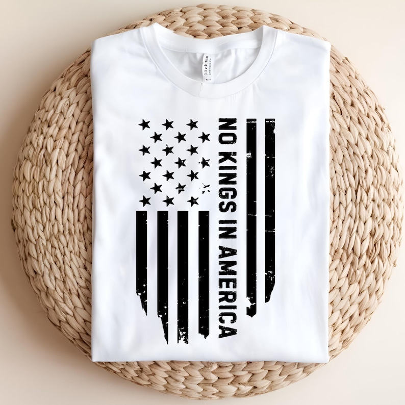 May include: White t-shirt featuring a distressed black graphic of the American flag with the text "NO KINGS IN AMERICA". The shirt is made of a soft material and is folded on a woven surface.