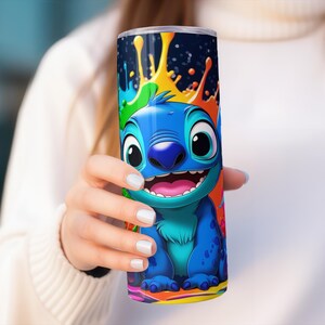 Puede incluir: Un vaso azul con un personaje de dibujos animados sonriendo ampliamente, sobre un dise&ntilde;o de salpicaduras colorido. El personaje es azul con la boca rosa. El fondo presenta salpicaduras verdes, naranjas y amarillas, creando un dise&ntilde;o vibrante.