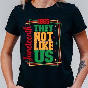 May include: Black t-shirt featuring a vibrant graphic. The design displays the text "1865 THEY NOT LIKE US" in green, yellow, and black, with the word "Juneteenth" in red on the left. The design is framed with red, green, and yellow lines.