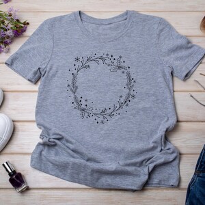 May include: A heather gray t-shirt featuring a black wreath design. The wreath is composed of sprigs, holly, and snowflakes. The t-shirt is made of a soft material and has short sleeves and a crew neck.
