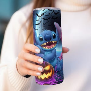 Pode incluir: Um copo azul com um design de Halloween com Stitch, uma ab&oacute;bora e morcegos contra um c&eacute;u iluminado pela lua. O copo &eacute; segurado por uma pessoa que usa um su&eacute;ter branco.
