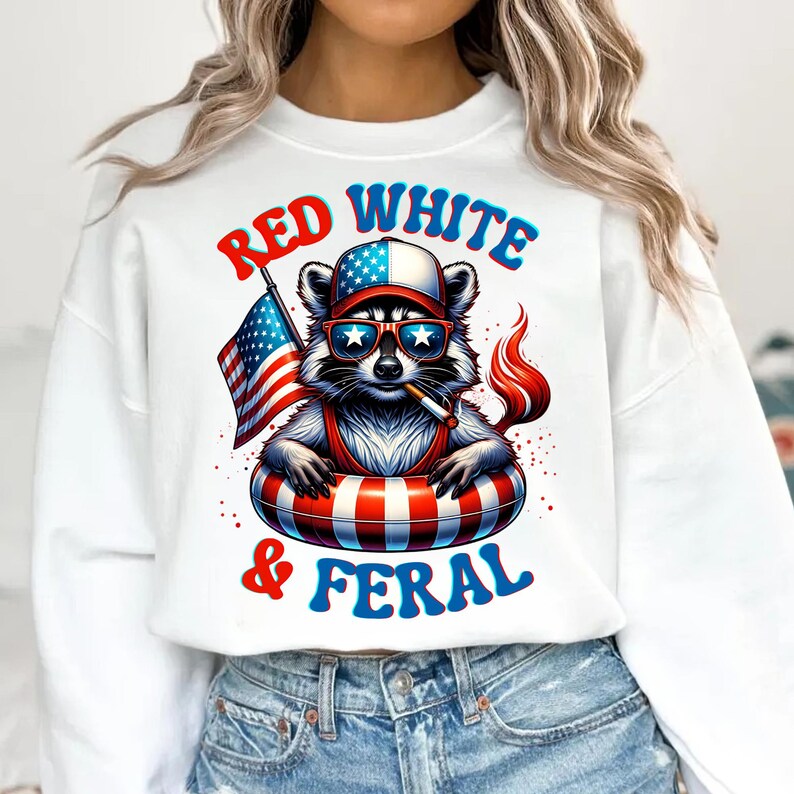 May include: White sweatshirt with a raccoon graphic wearing sunglasses, a hat, and an inner tube, holding a cigarette and an American flag. The text "Red White & Feral" is arched above and below the raccoon illustration.