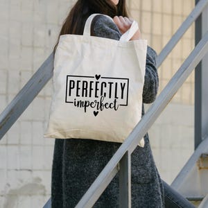 May include: A white canvas tote bag with black text that reads "Perfectly imperfect" with a heart symbol on either side of the text. The bag is being held by a person.