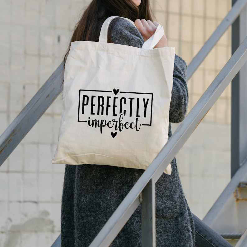 May include: A white canvas tote bag with black text that reads "Perfectly imperfect" with a heart symbol on either side of the text. The bag is being held by a person.
