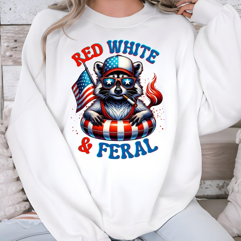May include: White sweatshirt with a raccoon graphic wearing sunglasses, a hat, and a red, white, and blue inner tube. The raccoon holds an American flag and a cigarette. The text "Red White & Feral" is displayed in red and blue.