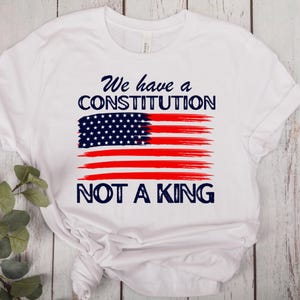 May include: White t-shirt featuring a stylized American flag design. The flag is composed of a blue field of stars and red stripes, with the text "We have a CONSTITUTION NOT A KING" in navy blue.