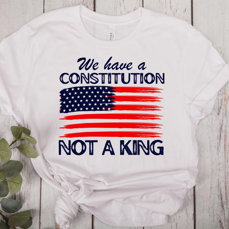 May include: White t-shirt featuring a stylized American flag design. The flag is composed of a blue field of stars and red stripes, with the text "We have a CONSTITUTION NOT A KING" in navy blue.