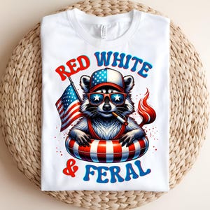 May include: White t-shirt with a raccoon graphic wearing sunglasses, a patriotic hat, and a red and white striped inner tube. The raccoon holds an American flag and a cigarette. The text on the shirt reads "RED WHITE & FERAL".
