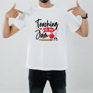 Op de afbeelding: Wit T-shirt met de tekst "Teaching is my Jam" in zwart schrift. Het ontwerp bevat een rode appel, een potlood, een schaar en andere schoolgerelateerde afbeeldingen. Het T-shirt heeft een ronde hals.