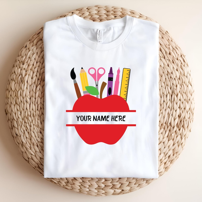 Op de afbeelding: Wit t-shirt met een rood appelontwerp. De appel heeft een ruimte voor personalisatie en is gevuld met schoolbenodigdheden zoals een penseel, potlood, schaar, kleurpotlood, liniaal en pennen. De tekst "YOUR NAME HERE" wordt weergegeven.