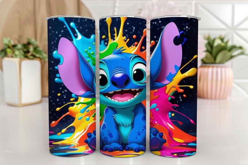 Puede incluir: Tres vasos de acero inoxidable con un personaje de dibujos animados azul, orejas grandes y una amplia sonrisa, rodeados de salpicaduras de pintura coloridas. El fondo es azul oscuro con motas blancas. Los vasos son cil&iacute;ndricos.