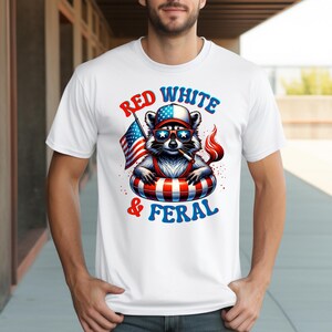 May include: White t-shirt with a cartoon raccoon wearing sunglasses, a hat, and an inner tube, holding an American flag and a lit cigarette. The text on the shirt reads "Red White & Feral" in red and blue.