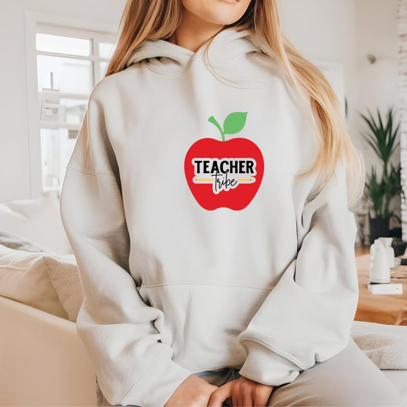 Op de afbeelding: Een witte hoodie met een rode appelafbeelding en een groen blad. Het appelontwerp bevat de woorden "Teacher Tribe" in zwart en wit. De hoodie is gemaakt van een zacht materiaal.