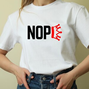 May include: White t-shirt with the word "NOPE" in black block letters and a red crown graphic. The shirt is made of a soft material and has a crew neck. The model is wearing blue jeans.