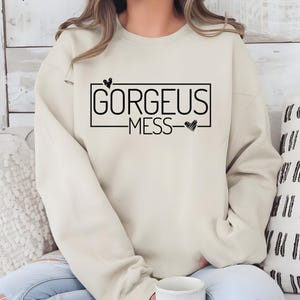 May include: A cream coloured sweatshirt with the text "GORGEOUS MESS" in a black rectangle with a small heart above and below the text.