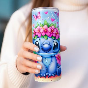 May include: A blue tumbler featuring a cartoon character with a floral crown and butterflies on a light blue background. The character has large eyes and a friendly expression. Pink flowers and hearts decorate the tumbler.