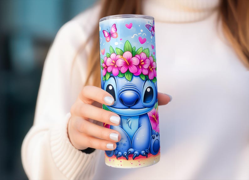 May include: A blue tumbler featuring a cartoon character with a floral crown and butterflies on a light blue background. The character has large eyes and a friendly expression. Pink flowers and hearts decorate the tumbler.