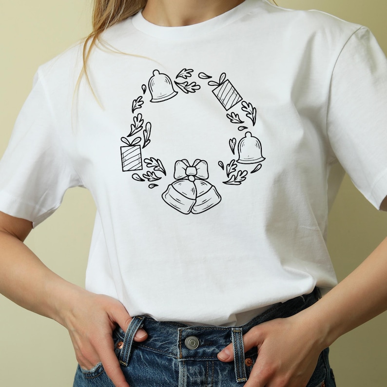 May include: White t-shirt featuring a black line art design of a wreath. The wreath includes bells, gift boxes, and leaves. The design is centered on the front of the shirt, creating a festive and decorative look.