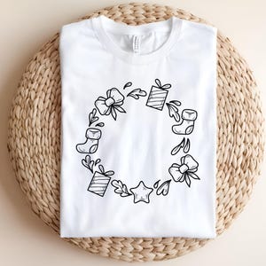 May include: White t-shirt featuring a black line drawing of a Christmas wreath. The wreath design includes bows, gift boxes, stockings, leaves, and a star. The shirt is displayed on a woven, beige surface.