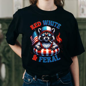 May include: Black t-shirt with a raccoon graphic wearing sunglasses, a hat, and a life preserver, holding an American flag and a cigarette. The text "RED WHITE & FERAL" is in red and blue.