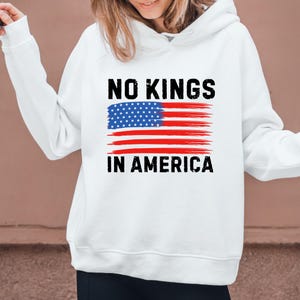 May include: White hoodie with the text "NO KINGS IN AMERICA" in black, above a stylized American flag design. The flag features a blue field of stars and red and white stripes, conveying a patriotic theme.