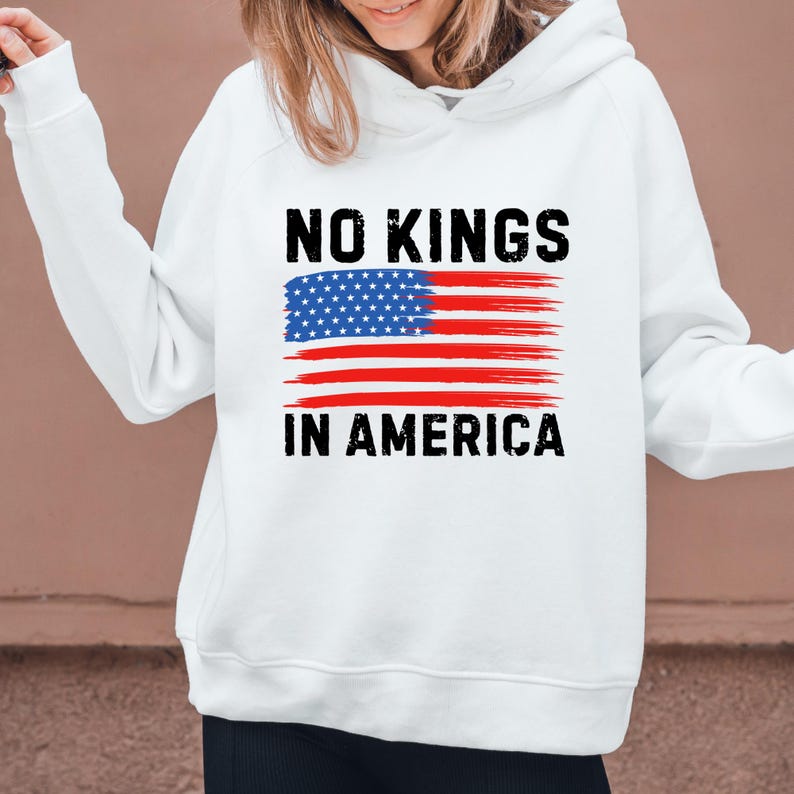 May include: White hoodie with the text "NO KINGS IN AMERICA" in black, above a stylized American flag design. The flag features a blue field of stars and red and white stripes, conveying a patriotic theme.