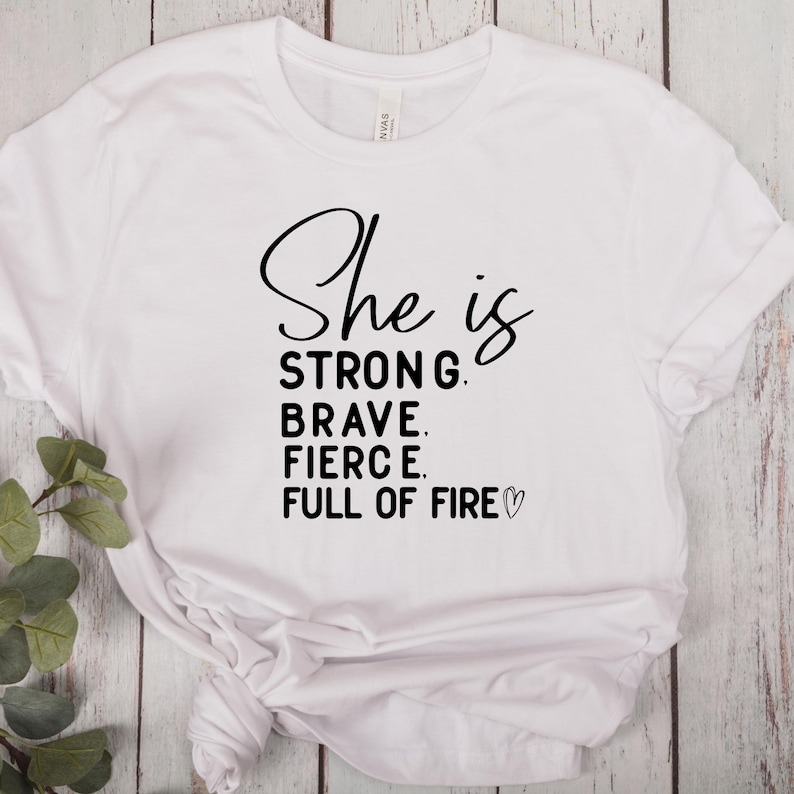 May include: White t-shirt with black text that reads "She is strong, brave, fierce, full of fire" with a small heart symbol.