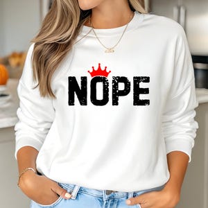 May include: White sweatshirt with the word "NOPE" in black, distressed font, topped with a red crown graphic. The sweatshirt is a casual style, suitable for everyday wear.