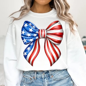 May include: White sweatshirt featuring a large bow design in red, white, and blue. The bow is styled to resemble the American flag, with blue sections adorned with white stars and red and white stripes.