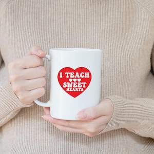 Op de afbeelding: Witte keramische mok met een rood hartontwerp. Het hart bevat de woorden "I TEACH SWEET HEARTS" in witte letters. De mok wordt vastgehouden door een persoon die een beige trui draagt.