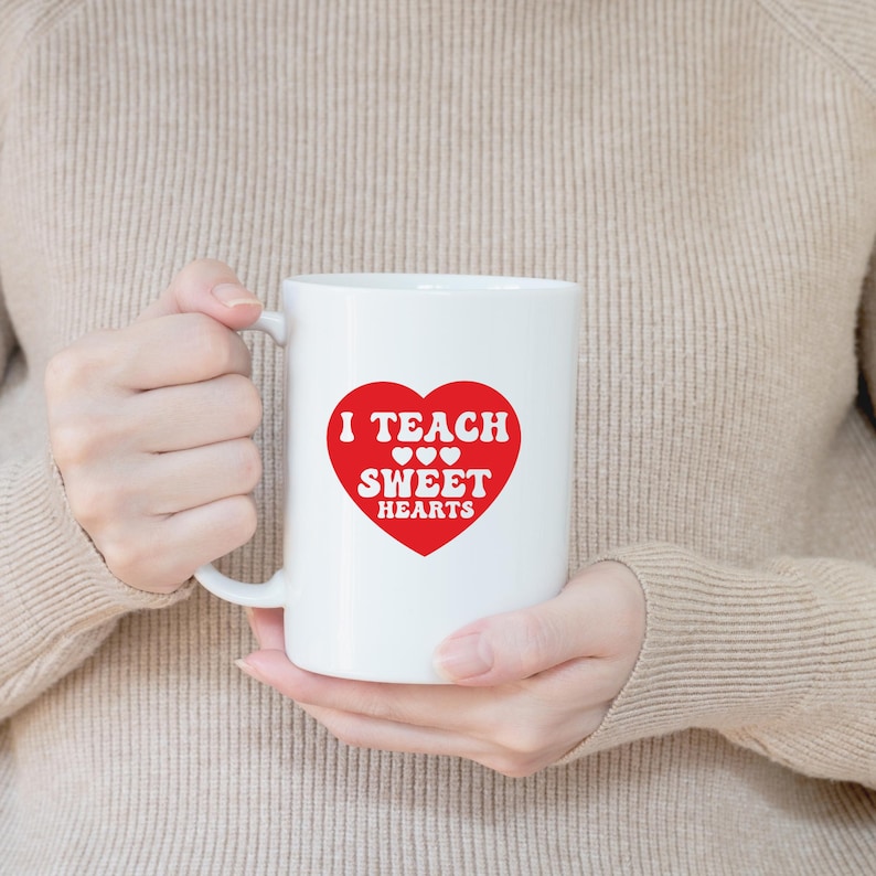 Op de afbeelding: Witte keramische mok met een rood hartontwerp. Het hart bevat de woorden "I TEACH SWEET HEARTS" in witte letters. De mok wordt vastgehouden door een persoon die een beige trui draagt.