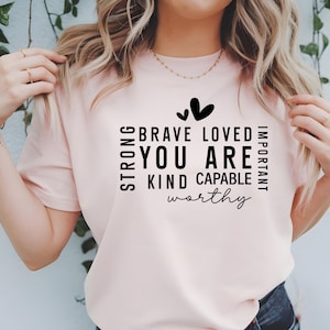 May include: A pink t-shirt with a black graphic that reads "Strong, Brave, Loved, Important, You Are, Kind, Capable, Worthy" with two small hearts above the text.