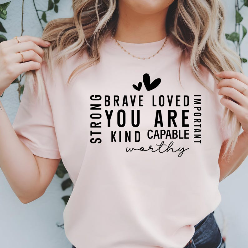 May include: A pink t-shirt with a black graphic that reads "Strong, Brave, Loved, Important, You Are, Kind, Capable, Worthy" with two small hearts above the text.